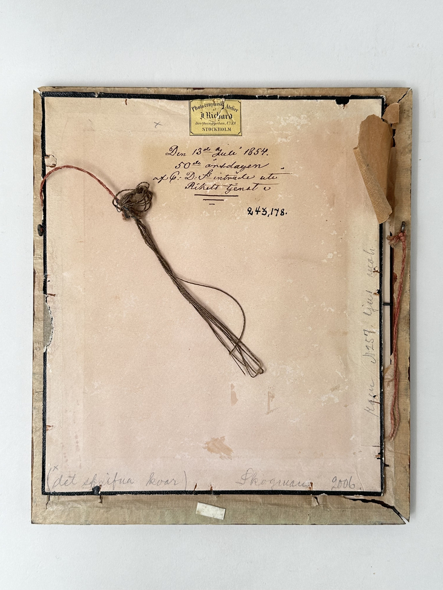 Fotografi i fyrkantig ram av trä med svarta hörn. Fotografiet föreställandes en sittande man i 70-årsåldern, uppklädd med ett ordenstecken på kavajen och käpp i handen. Naturfärgad passepartout med oval öppning för fotografiet, rand i guld och vitt. Baksidan i papp. 
Ramen trasig i hörnen, motivet ligger löst. Glaset i ramen ojämnt klippt upptill. 