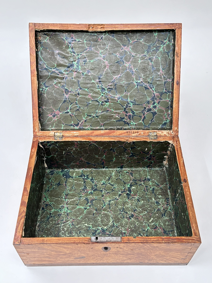 Skrin i trä klädd i två olika faner. Lockets ram, kring nyckelhål samt lådans nederkanter i mörk faner; i övrigt ljusare faner. Låset saknar ett beslag samt nyckel. Locket fäst i lådan med små platta gångjärn. Insidan klädd i marmorerat papper i grönaktiga toner. 