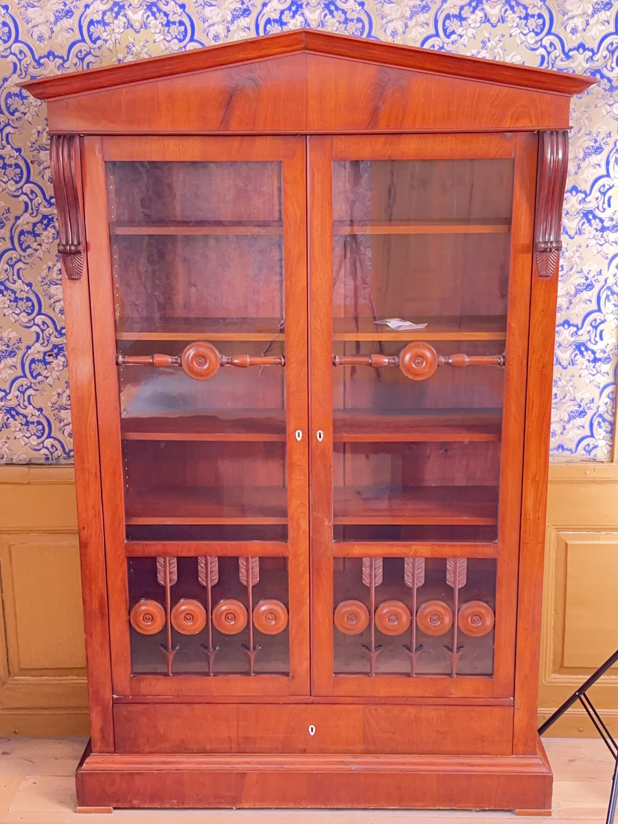 Bokskåp i mahogny med glasdörrar och låda nederst. Gavelformat överstycke med plats för fyra hörnkrön. Två krön har blomornament på två sidor, de andra två krönen har endast ornament på den ena sidan. Fem hyllplan, 20 platta hyllbärare i metall. Hålen för hyllbärarna klädda i mässing. Glasdörrarna med snidade dekorationer i form av bland annat cirklar och pilar. Dekorativa konsoler på bägge sidor dörrarna. Nyckelbrickor till dörrar och låda i ben.