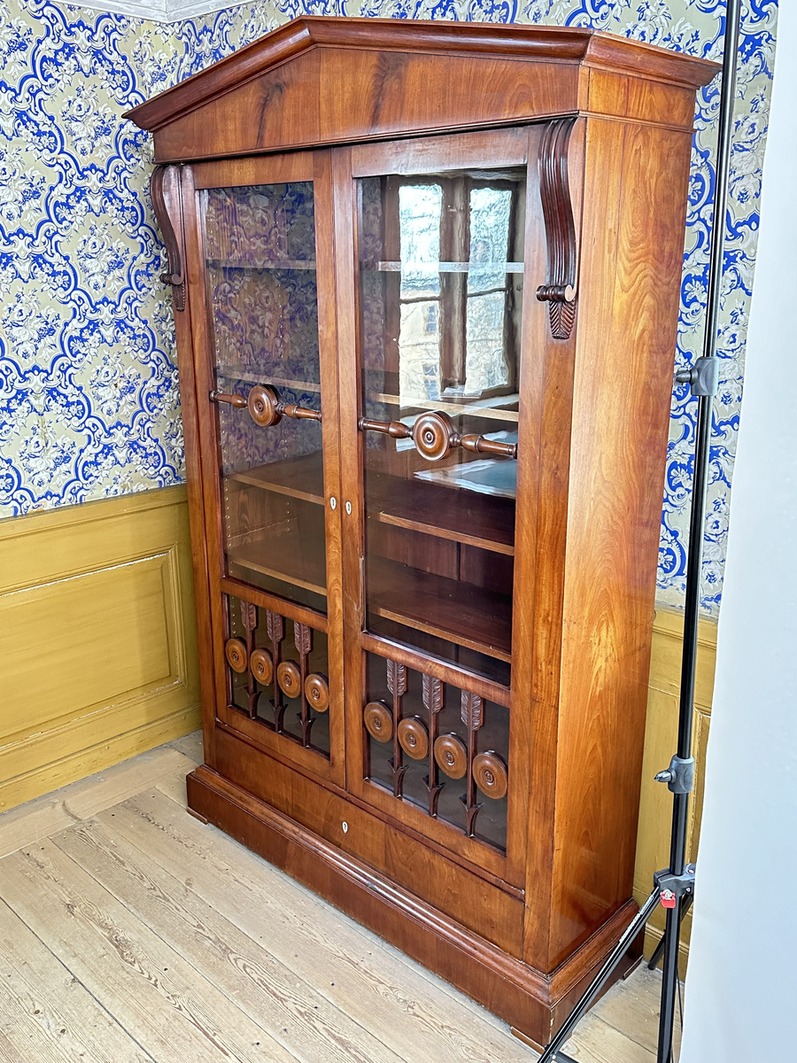 Bokskåp i mahogny med glasdörrar och låda nederst. Gavelformat överstycke med plats för fyra hörnkrön. Två krön har blomornament på två sidor, de andra två krönen har endast ornament på den ena sidan. Fem hyllplan, 20 platta hyllbärare i metall. Hålen för hyllbärarna klädda i mässing. Glasdörrarna med snidade dekorationer i form av bland annat cirklar och pilar. Dekorativa konsoler på bägge sidor dörrarna. Nyckelbrickor till dörrar och låda i ben.