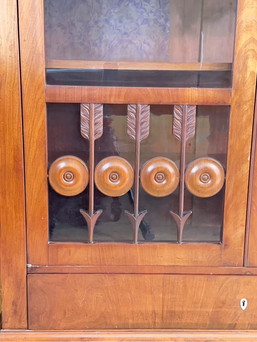 Bokskåp i mahogny med glasdörrar och låda nederst. Gavelformat överstycke med plats för fyra hörnkrön. Två krön har blomornament på två sidor, de andra två krönen har endast ornament på den ena sidan. Fem hyllplan, 20 platta hyllbärare i metall. Hålen för hyllbärarna klädda i mässing. Glasdörrarna med snidade dekorationer i form av bland annat cirklar och pilar. Dekorativa konsoler på bägge sidor dörrarna. Nyckelbrickor till dörrar och låda i ben.