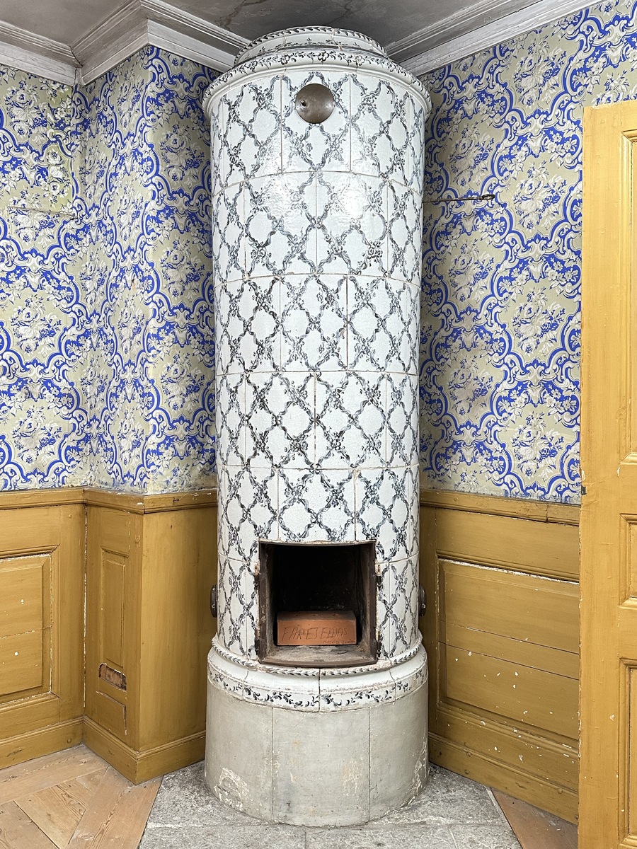 Kakelugn, rund. Klädd i kvadratiskt vitglaserat kakel, handmålat med korsande blomstergirlander i duvblått och mörkblåa detaljer. Slät sockel, gråkalkad. Fot- och översims med växlande konvex och konkav profilering samt liggande blomstergirlander. Två runda sotluckor i mässing på ugnens sidor ovanför sockeln, samt en på ugnens topp. Spjällarm av järn med ringformat handtag.