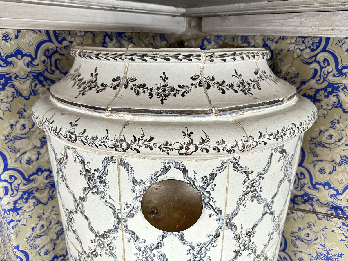 Kakelugn, rund. Klädd i kvadratiskt vitglaserat kakel, handmålat med korsande blomstergirlander i duvblått och mörkblåa detaljer. Slät sockel, gråkalkad. Fot- och översims med växlande konvex och konkav profilering samt liggande blomstergirlander. Två runda sotluckor i mässing på ugnens sidor ovanför sockeln, samt en på ugnens topp. Spjällarm av järn med ringformat handtag.