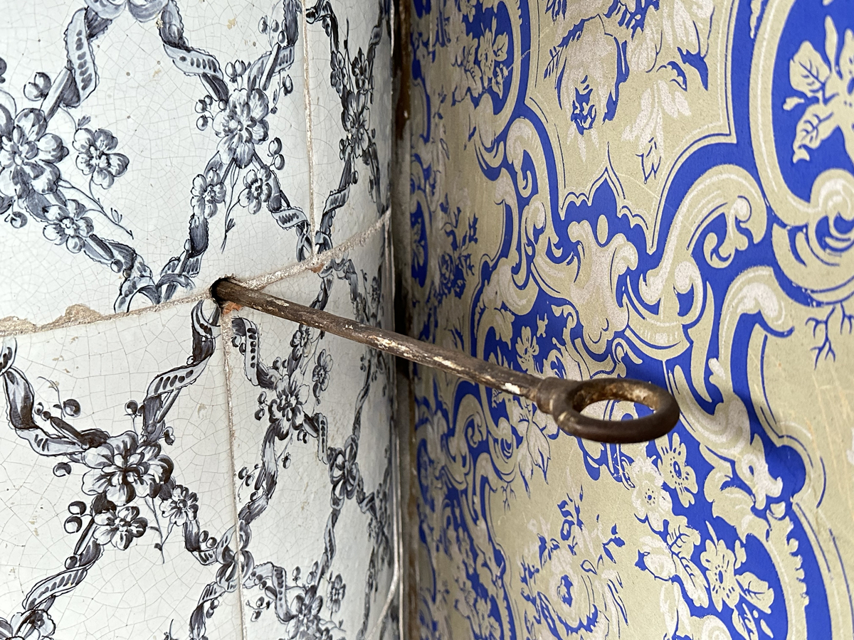 Kakelugn, rund. Klädd i kvadratiskt vitglaserat kakel, handmålat med korsande blomstergirlander i duvblått och mörkblåa detaljer. Slät sockel, gråkalkad. Fot- och översims med växlande konvex och konkav profilering samt liggande blomstergirlander. Två runda sotluckor i mässing på ugnens sidor ovanför sockeln, samt en på ugnens topp. Spjällarm av järn med ringformat handtag.