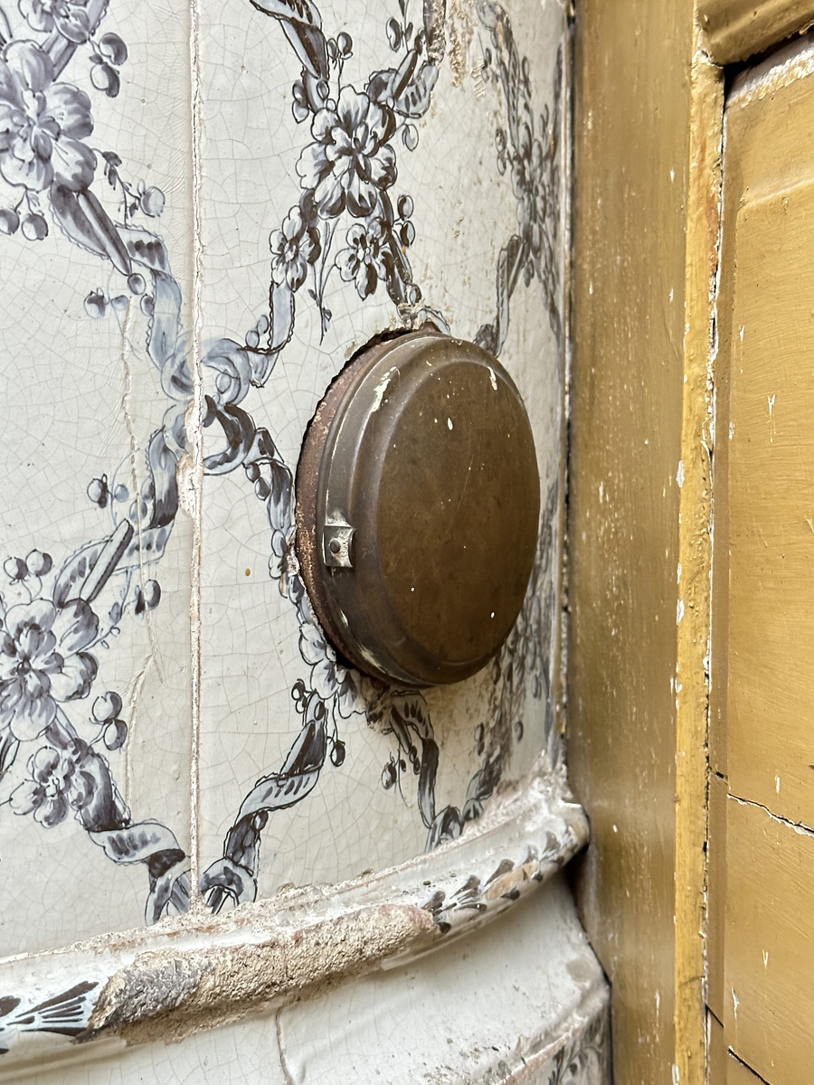 Kakelugn, rund. Klädd i kvadratiskt vitglaserat kakel, handmålat med korsande blomstergirlander i duvblått och mörkblåa detaljer. Slät sockel, gråkalkad. Fot- och översims med växlande konvex och konkav profilering samt liggande blomstergirlander. Två runda sotluckor i mässing på ugnens sidor ovanför sockeln, samt en på ugnens topp. Spjällarm av järn med ringformat handtag.