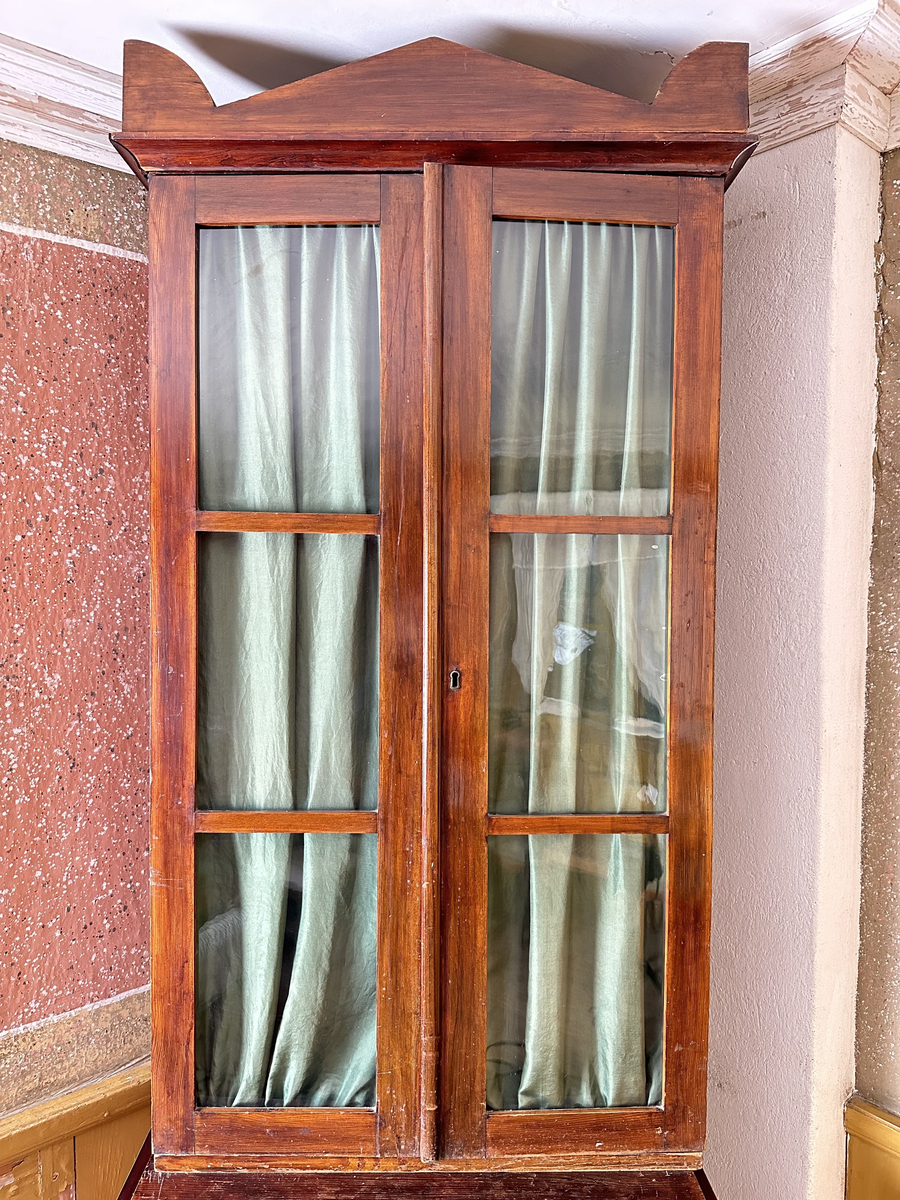 Skåp, överskåp till bokskåp. Av furu, mörkt betsad. Dubbeldörrar, fyllda med tre glasrutor vardera. Glaset munblåst med ojämnheter och små, fina lusor. Tygvåder av skogsgrönt siden hängandes för glasrutorna på insidan dörrarna. Infälld låskista och mässingsomfattat nyckelhål i den högra dörren. Juster- och löstagbara hyllplan, fyra stycken. Överst på skåpet ett aningen utkragande, enkelt krön.