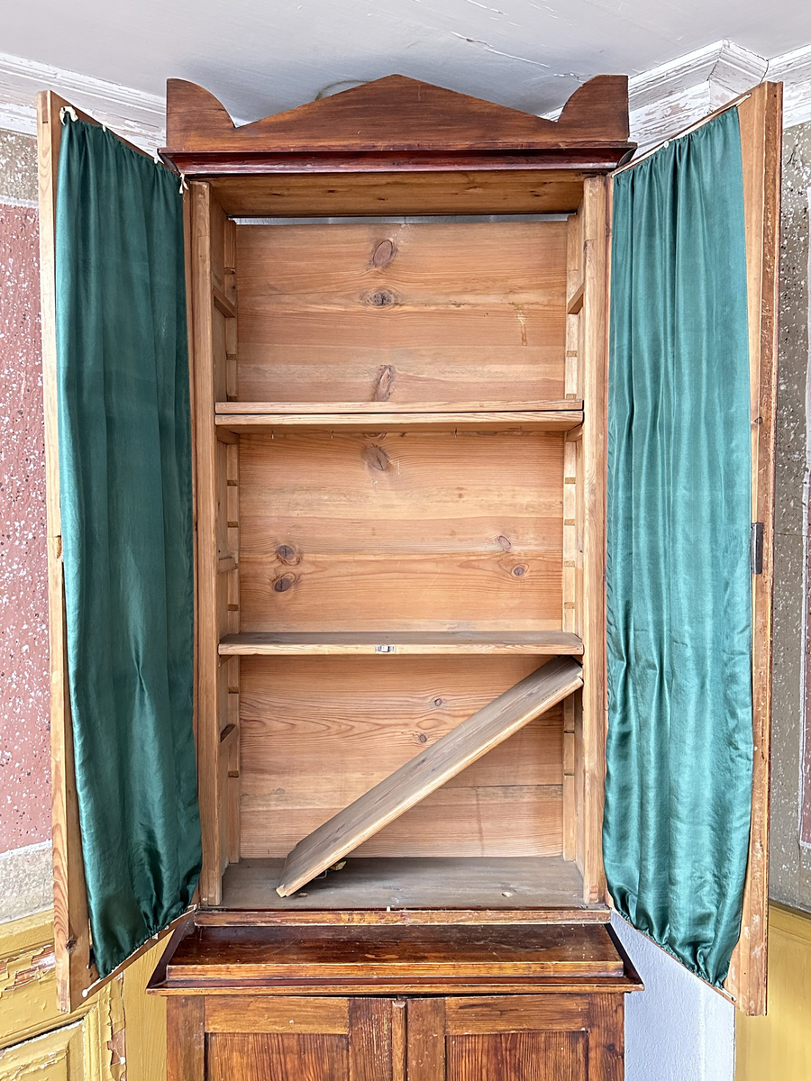 Skåp, överskåp till bokskåp. Av furu, mörkt betsad. Dubbeldörrar, fyllda med tre glasrutor vardera. Glaset munblåst med ojämnheter och små, fina lusor. Tygvåder av skogsgrönt siden hängandes för glasrutorna på insidan dörrarna. Infälld låskista och mässingsomfattat nyckelhål i den högra dörren. Juster- och löstagbara hyllplan, fyra stycken. Överst på skåpet ett aningen utkragande, enkelt krön.