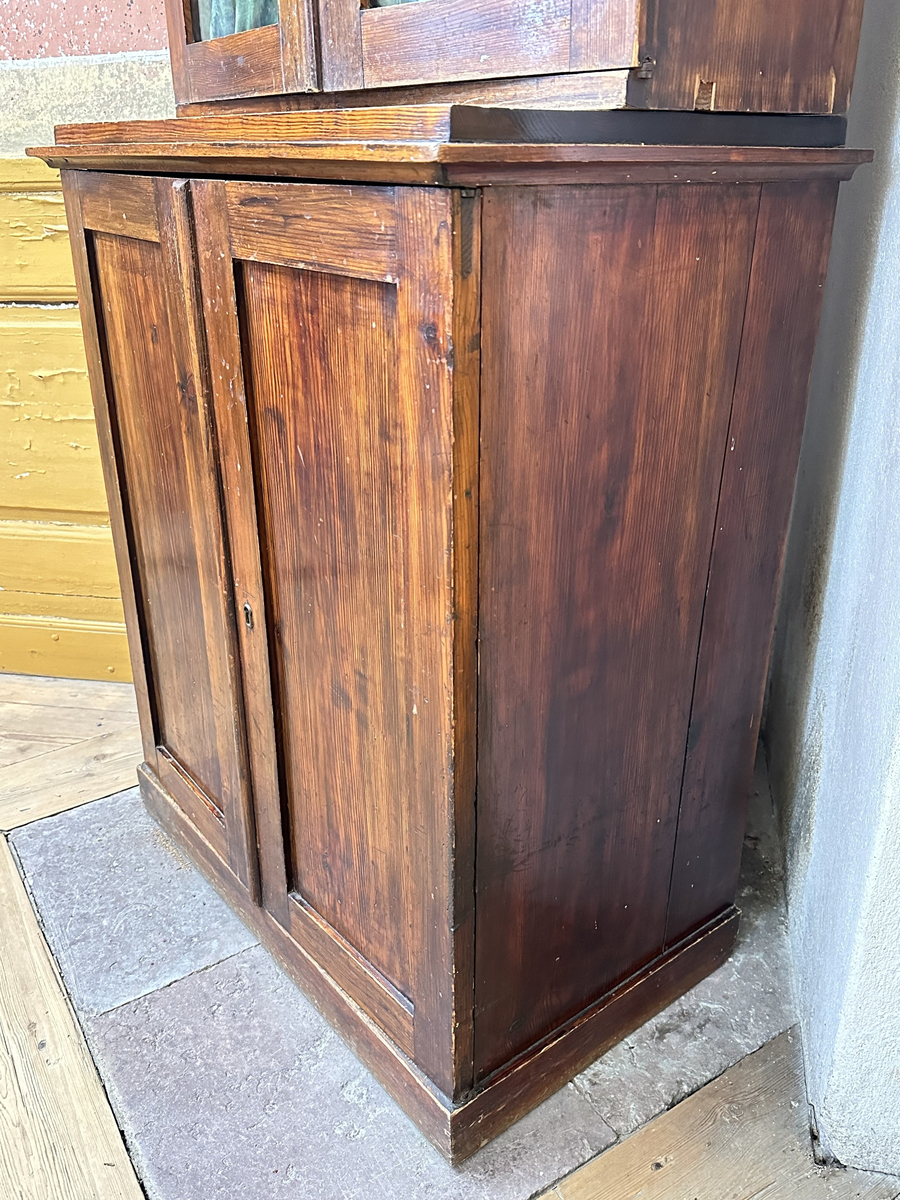 Skåp, underdel av bokskåp. Av furu, mörkt betsad. Ståendes på en slät sockel. Dubbeldörrar med en fyllning vardera, samt infälld låskista och mässingsomfattat nyckelhål i den högra dörren. Toppskivan profilerad i kanterna, med ovanliggande platå. Juster- och löstagbart hyllplan, två stycken.