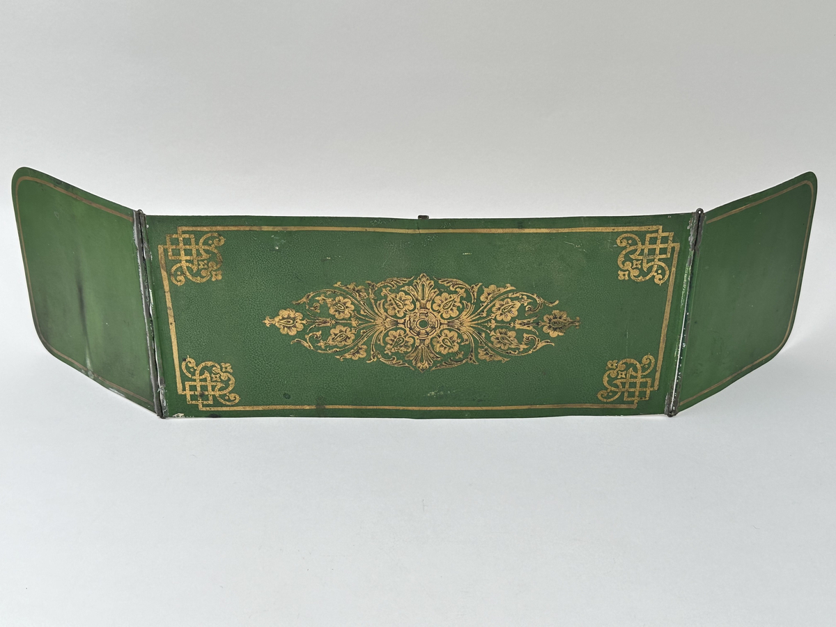 Skärm till ljusstake, i lackerad metallplåt. En rektangulär skärm, med två vinklingsbara vingar på vardera sida. Framsida grönlackerad med guldornamentik. Baksida vitlackerad, med en del krackelyr och nötning på lacken. Pålött fäste, centrerat på skärmens baksida.