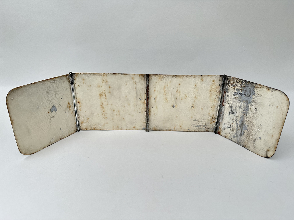 Skärm till ljusstake, i lackerad metallplåt. En rektangulär skärm, med två vinklingsbara vingar på vardera sida. Framsida grönlackerad med guldornamentik. Baksida vitlackerad, med en del krackelyr och nötning på lacken. Pålött fäste, centrerat på skärmens baksida.