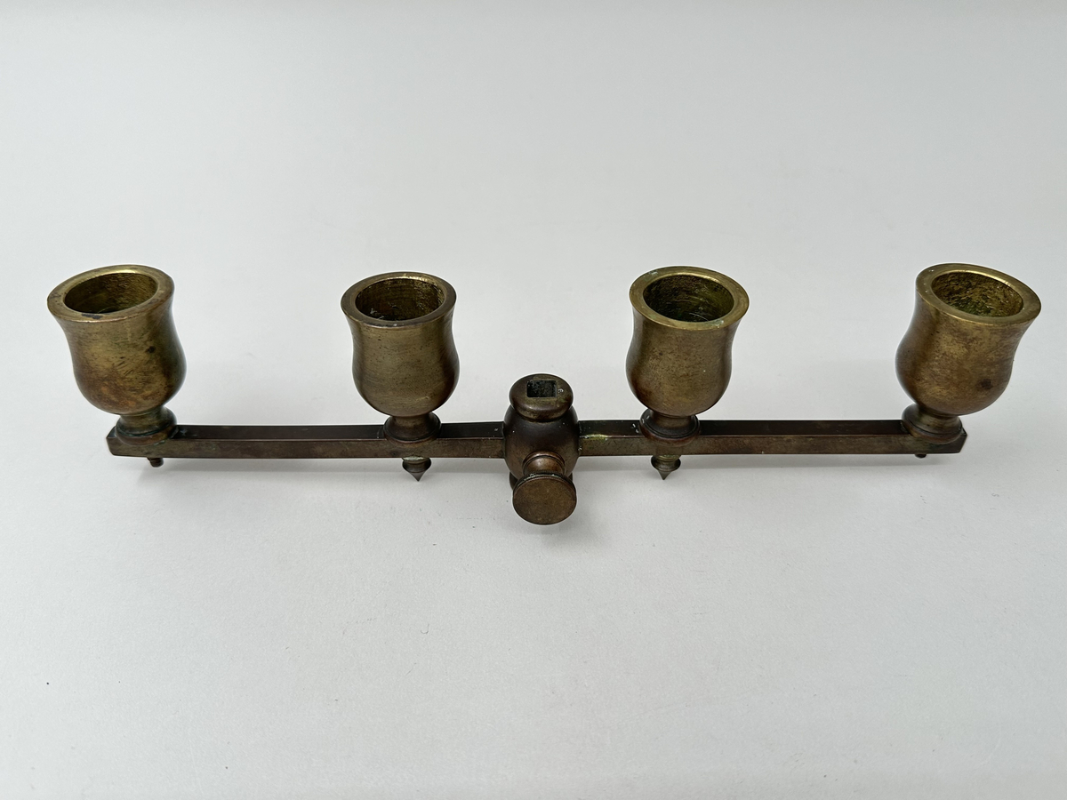 Ställbar, rak arm för kronljus av brunpatinerad mässing. Fyra ljuspipor, tulpanformade, fästa med skruv som tränger ut genom armens undersida. De två innersta pipornas skruvändar dolda med spetsiga dekorativa nitar. Fäste i mjuk form, med två vågräta indragningar och centrerat fyrkantigt hål. Rund skruv med räfflade kanter för att fästa armen i läge på ståndaren.