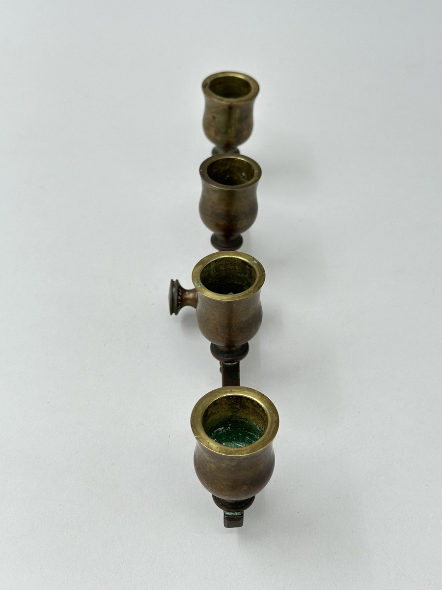 Ställbar, rak arm för kronljus av brunpatinerad mässing. Fyra ljuspipor, tulpanformade, fästa med skruv som tränger ut genom armens undersida. De två innersta pipornas skruvändar dolda med spetsiga dekorativa nitar. Fäste i mjuk form, med två vågräta indragningar och centrerat fyrkantigt hål. Rund skruv med räfflade kanter för att fästa armen i läge på ståndaren.