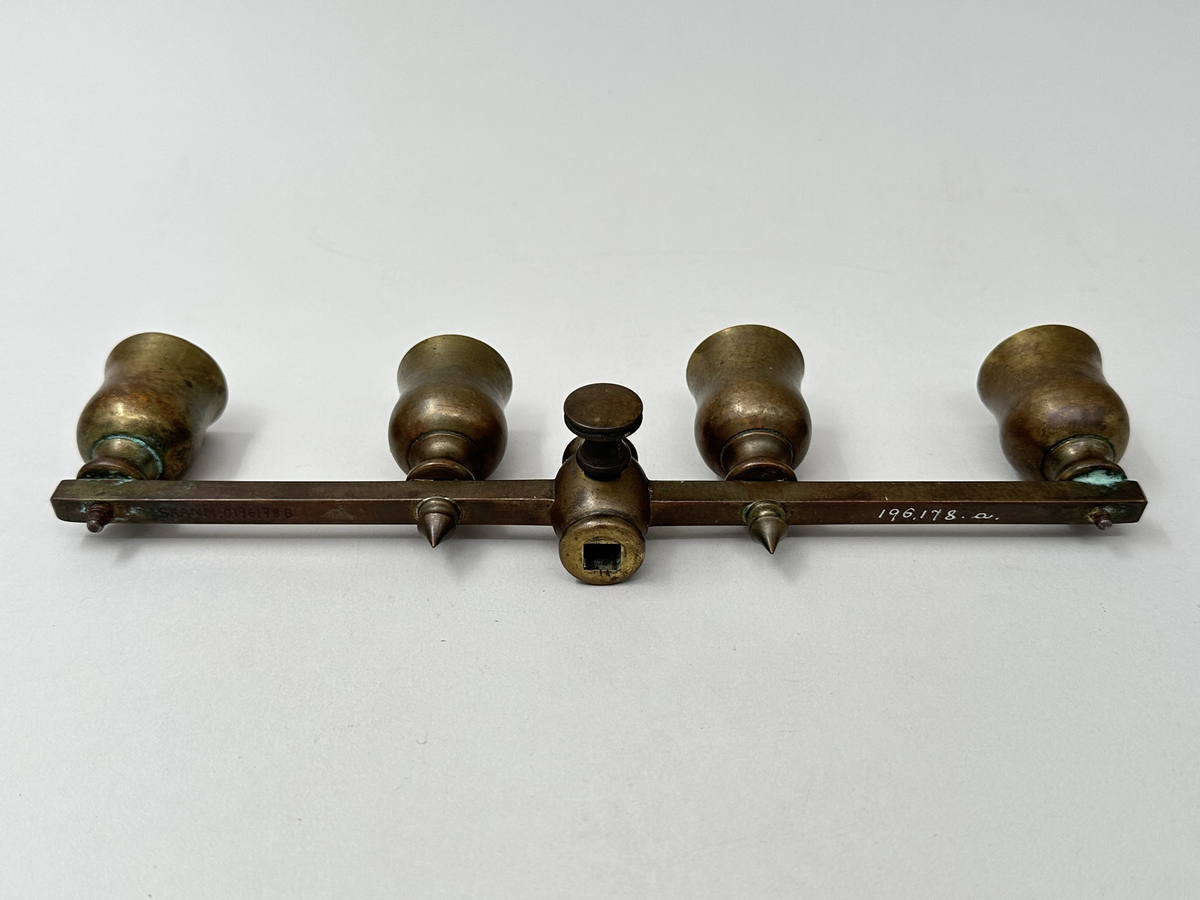 Ställbar, rak arm för kronljus av brunpatinerad mässing. Fyra ljuspipor, tulpanformade, fästa med skruv som tränger ut genom armens undersida. De två innersta pipornas skruvändar dolda med spetsiga dekorativa nitar. Fäste i mjuk form, med två vågräta indragningar och centrerat fyrkantigt hål. Rund skruv med räfflade kanter för att fästa armen i läge på ståndaren.