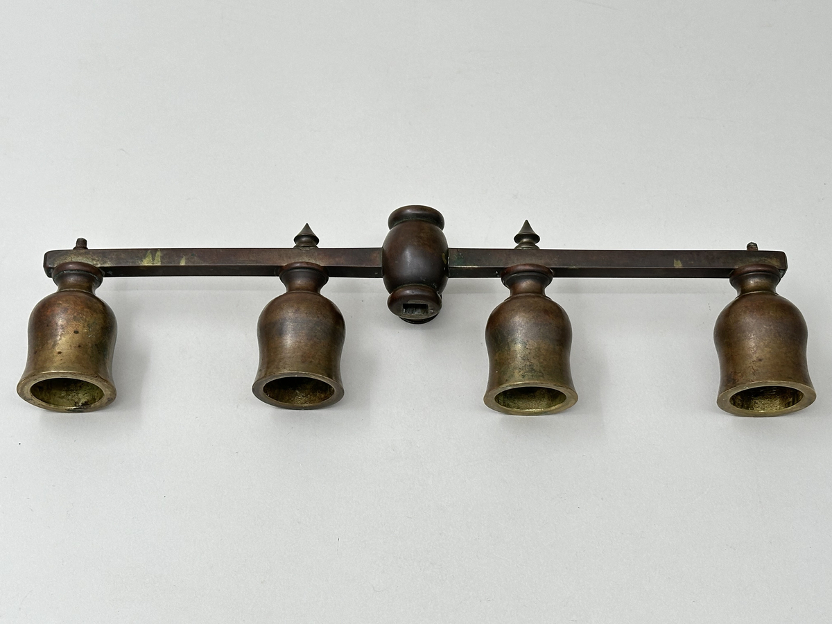 Ställbar, rak arm för kronljus av brunpatinerad mässing. Fyra ljuspipor, tulpanformade, fästa med skruv som tränger ut genom armens undersida. De två innersta pipornas skruvändar dolda med spetsiga dekorativa nitar. Fäste i mjuk form, med två vågräta indragningar och centrerat fyrkantigt hål. Rund skruv med räfflade kanter för att fästa armen i läge på ståndaren.