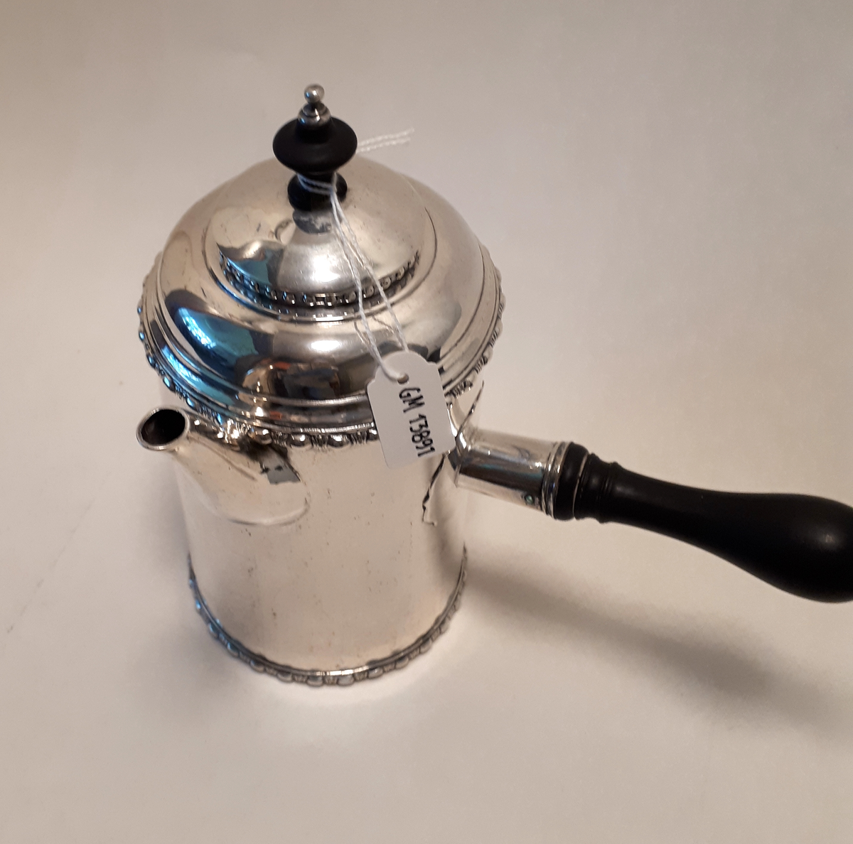 Kaffekanna i silver. Gustaviansk, cylindrisk med raka sidor, drivna bårder upptill och nertill, kort lätt böjd pip, kupigt lock med svart träknopp med silvertopp. Rakt utstående svarvat trähandtag. Graverat i botten "N:G:H, 1765" samt viktangivelsen "4 1 1/1 lod".