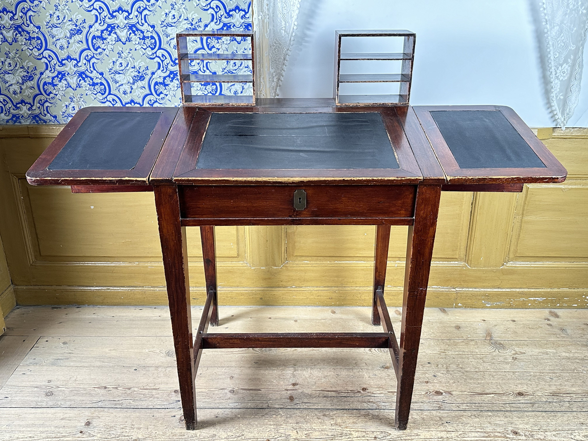 Pulpet, av mörkt betsat trä. Skrivyta med två fällbara sidoskivor, klädda med svart pergamoid. Sidoskivorna hålls uppe med utfällbara utanpåliggande delar av sargen, rakt sinkade, med skålade handtag. Intill dessa ytterligare utfällbara bitar som blir avstånd mot vägg. Sarg med låsbar förvaring, åtkomlig via lyft av skrivyta, nyckelskylt av mässing framtill. Två små hyllor på pulpetens krön med tre fack vardera, laxstjärtssinkade. Fyra raka, fyrkantiga ben med avsmalning nedåt som sammanbinds med ett H-stag.