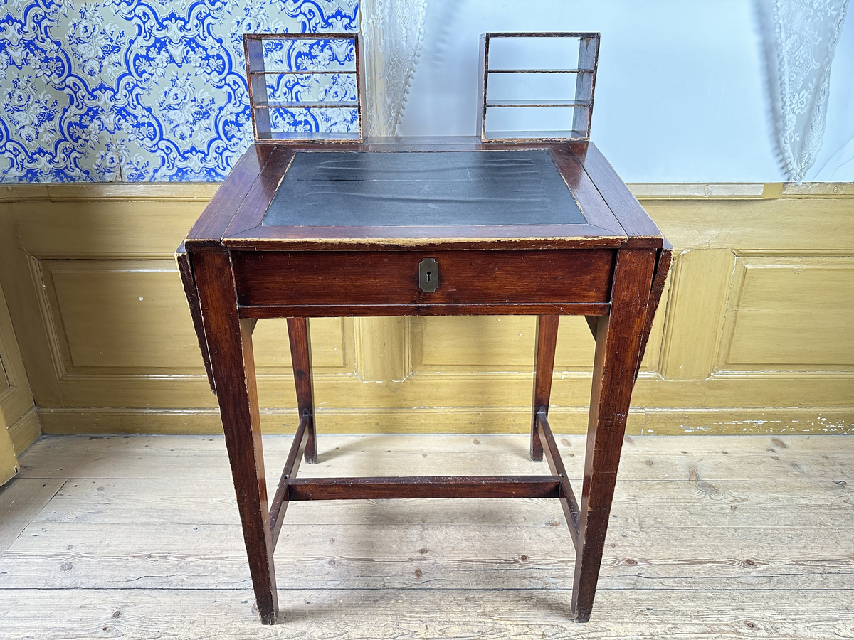 Pulpet, av mörkt betsat trä. Skrivyta med två fällbara sidoskivor, klädda med svart pergamoid. Sidoskivorna hålls uppe med utfällbara utanpåliggande delar av sargen, rakt sinkade, med skålade handtag. Intill dessa ytterligare utfällbara bitar som blir avstånd mot vägg. Sarg med låsbar förvaring, åtkomlig via lyft av skrivyta, nyckelskylt av mässing framtill. Två små hyllor på pulpetens krön med tre fack vardera, laxstjärtssinkade. Fyra raka, fyrkantiga ben med avsmalning nedåt som sammanbinds med ett H-stag.