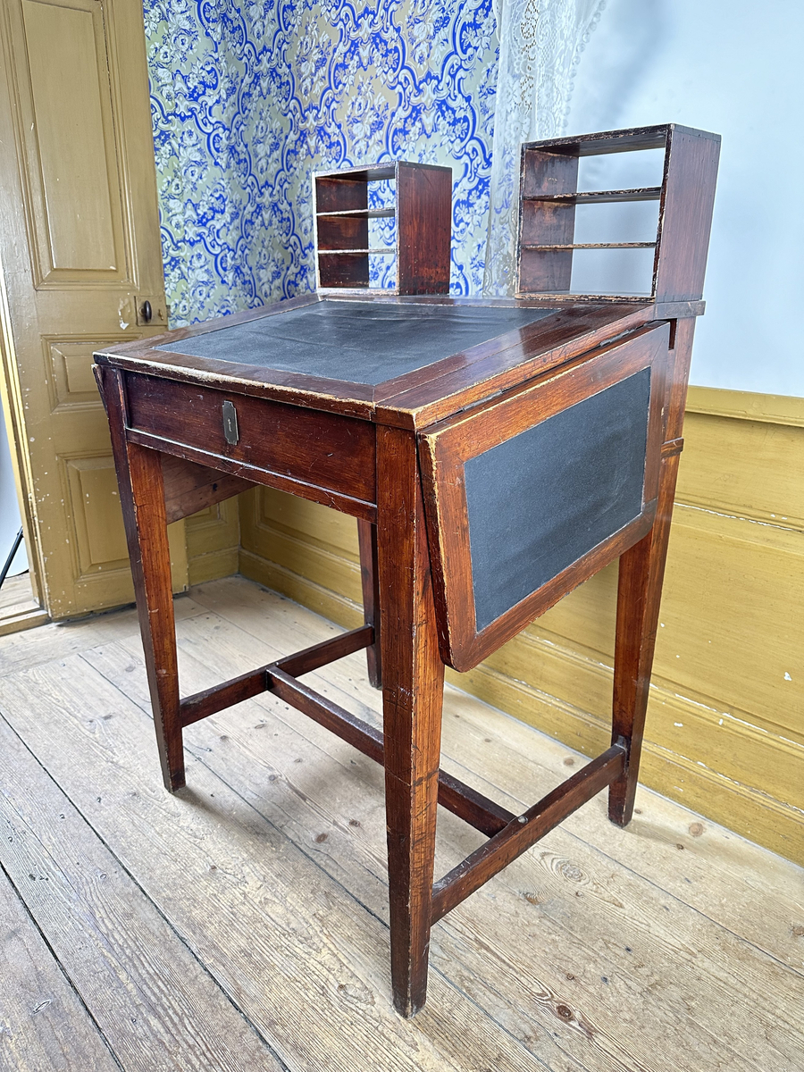 Pulpet, av mörkt betsat trä. Skrivyta med två fällbara sidoskivor, klädda med svart pergamoid. Sidoskivorna hålls uppe med utfällbara utanpåliggande delar av sargen, rakt sinkade, med skålade handtag. Intill dessa ytterligare utfällbara bitar som blir avstånd mot vägg. Sarg med låsbar förvaring, åtkomlig via lyft av skrivyta, nyckelskylt av mässing framtill. Två små hyllor på pulpetens krön med tre fack vardera, laxstjärtssinkade. Fyra raka, fyrkantiga ben med avsmalning nedåt som sammanbinds med ett H-stag.