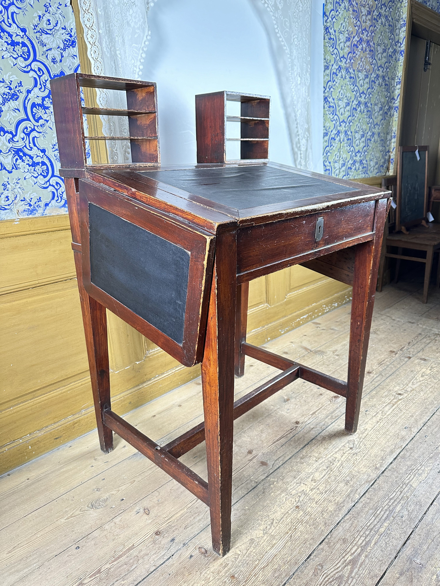 Pulpet, av mörkt betsat trä. Skrivyta med två fällbara sidoskivor, klädda med svart pergamoid. Sidoskivorna hålls uppe med utfällbara utanpåliggande delar av sargen, rakt sinkade, med skålade handtag. Intill dessa ytterligare utfällbara bitar som blir avstånd mot vägg. Sarg med låsbar förvaring, åtkomlig via lyft av skrivyta, nyckelskylt av mässing framtill. Två små hyllor på pulpetens krön med tre fack vardera, laxstjärtssinkade. Fyra raka, fyrkantiga ben med avsmalning nedåt som sammanbinds med ett H-stag.