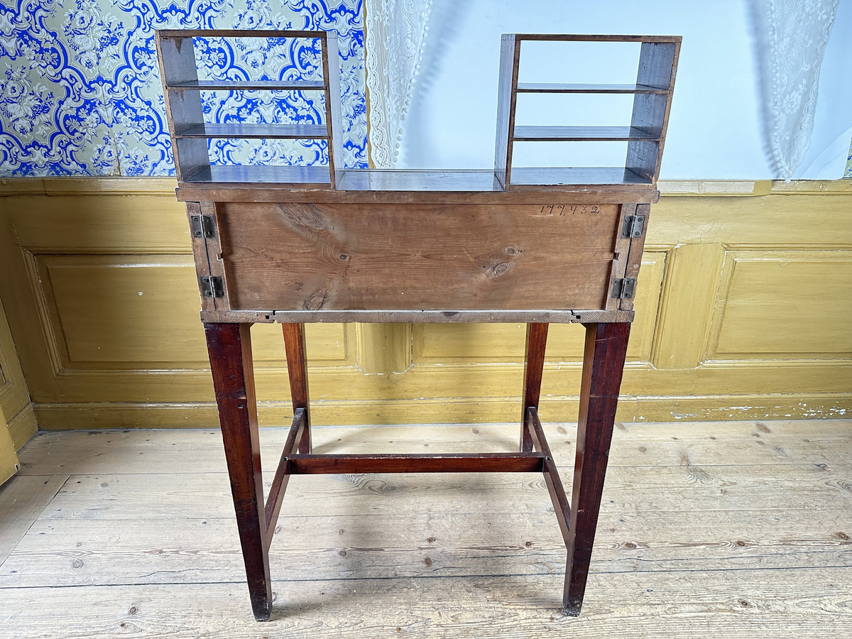 Pulpet, av mörkt betsat trä. Skrivyta med två fällbara sidoskivor, klädda med svart pergamoid. Sidoskivorna hålls uppe med utfällbara utanpåliggande delar av sargen, rakt sinkade, med skålade handtag. Intill dessa ytterligare utfällbara bitar som blir avstånd mot vägg. Sarg med låsbar förvaring, åtkomlig via lyft av skrivyta, nyckelskylt av mässing framtill. Två små hyllor på pulpetens krön med tre fack vardera, laxstjärtssinkade. Fyra raka, fyrkantiga ben med avsmalning nedåt som sammanbinds med ett H-stag.