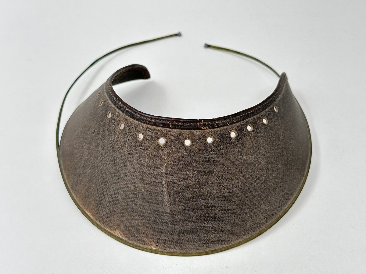 Ögonskärm, av vaxat klot med brun utsida och grön insida. Mot pannan kantat med läderband, samt perforerat med 13 hål. Skärmens nederkant fäst i bygel av mässing, med pålödda kuländar.
