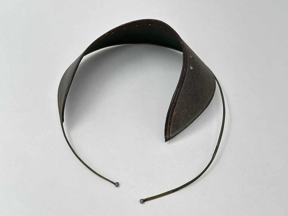 Ögonskärm, av vaxat klot med brun utsida och grön insida. Mot pannan kantat med läderband, samt perforerat med 13 hål. Skärmens nederkant fäst i bygel av mässing, med pålödda kuländar.