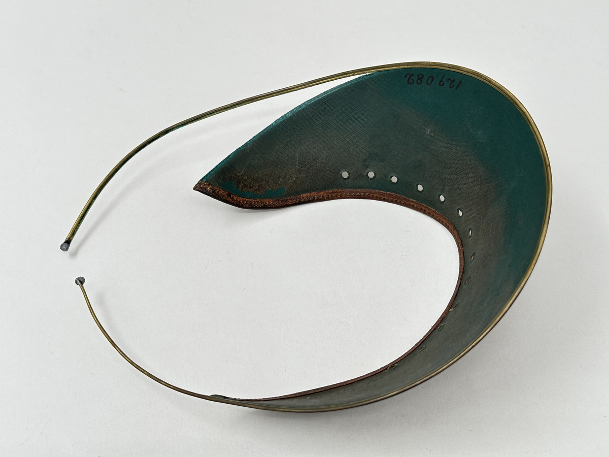 Ögonskärm, av vaxat klot med brun utsida och grön insida. Mot pannan kantat med läderband, samt perforerat med 13 hål. Skärmens nederkant fäst i bygel av mässing, med pålödda kuländar.