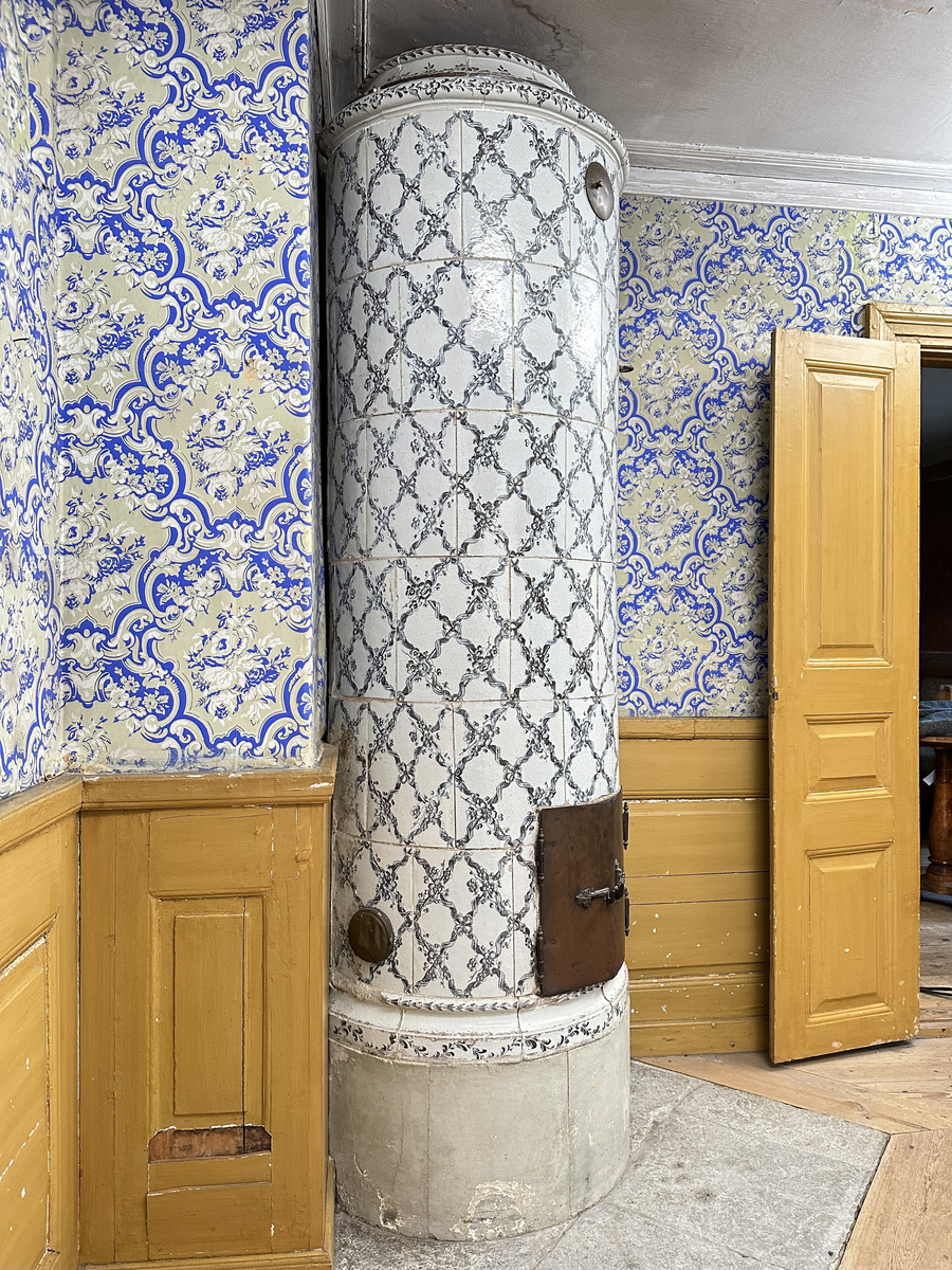 Kakelugn, rund. Klädd i kvadratiskt vitglaserat kakel, handmålat med korsande blomstergirlander i duvblått och mörkblåa detaljer. Slät sockel, gråkalkad. Fot- och översims med växlande konvex och konkav profilering samt liggande blomstergirlander. Ytterluckor i mässing med dekorativt utformade gångjärn samt klinka, i form av en sädeskärve med blommor. Innerluckor av järn, rostiga, med klinka samt två päronformade ventilluckor. Två runda sotluckor i mässing på ugnens sidor ovanför sockeln, samt en på ugnens topp. Spjällarm med ringformat handtag av järn.