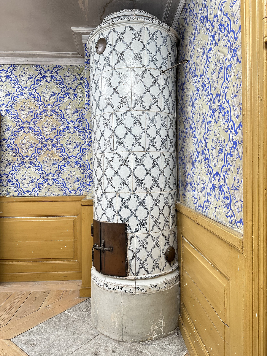 Kakelugn, rund. Klädd i kvadratiskt vitglaserat kakel, handmålat med korsande blomstergirlander i duvblått och mörkblåa detaljer. Slät sockel, gråkalkad. Fot- och översims med växlande konvex och konkav profilering samt liggande blomstergirlander. Ytterluckor i mässing med dekorativt utformade gångjärn samt klinka, i form av en sädeskärve med blommor. Innerluckor av järn, rostiga, med klinka samt två päronformade ventilluckor. Två runda sotluckor i mässing på ugnens sidor ovanför sockeln, samt en på ugnens topp. Spjällarm med ringformat handtag av järn.