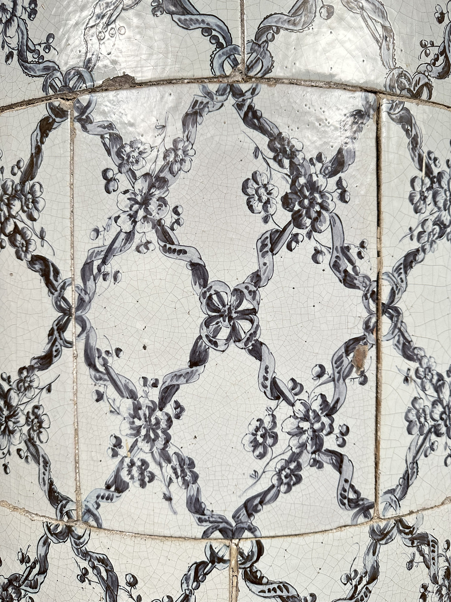 Kakelugn, rund. Klädd i kvadratiskt vitglaserat kakel, handmålat med korsande blomstergirlander i duvblått och mörkblåa detaljer. Slät sockel, gråkalkad. Fot- och översims med växlande konvex och konkav profilering samt liggande blomstergirlander. Ytterluckor i mässing med dekorativt utformade gångjärn samt klinka, i form av en sädeskärve med blommor. Innerluckor av järn, rostiga, med klinka samt två päronformade ventilluckor. Två runda sotluckor i mässing på ugnens sidor ovanför sockeln, samt en på ugnens topp. Spjällarm med ringformat handtag av järn.