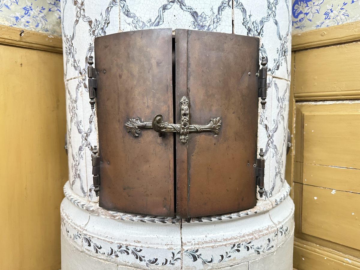 Kakelugn, rund. Klädd i kvadratiskt vitglaserat kakel, handmålat med korsande blomstergirlander i duvblått och mörkblåa detaljer. Slät sockel, gråkalkad. Fot- och översims med växlande konvex och konkav profilering samt liggande blomstergirlander. Ytterluckor i mässing med dekorativt utformade gångjärn samt klinka, i form av en sädeskärve med blommor. Innerluckor av järn, rostiga, med klinka samt två päronformade ventilluckor. Två runda sotluckor i mässing på ugnens sidor ovanför sockeln, samt en på ugnens topp. Spjällarm med ringformat handtag av järn.