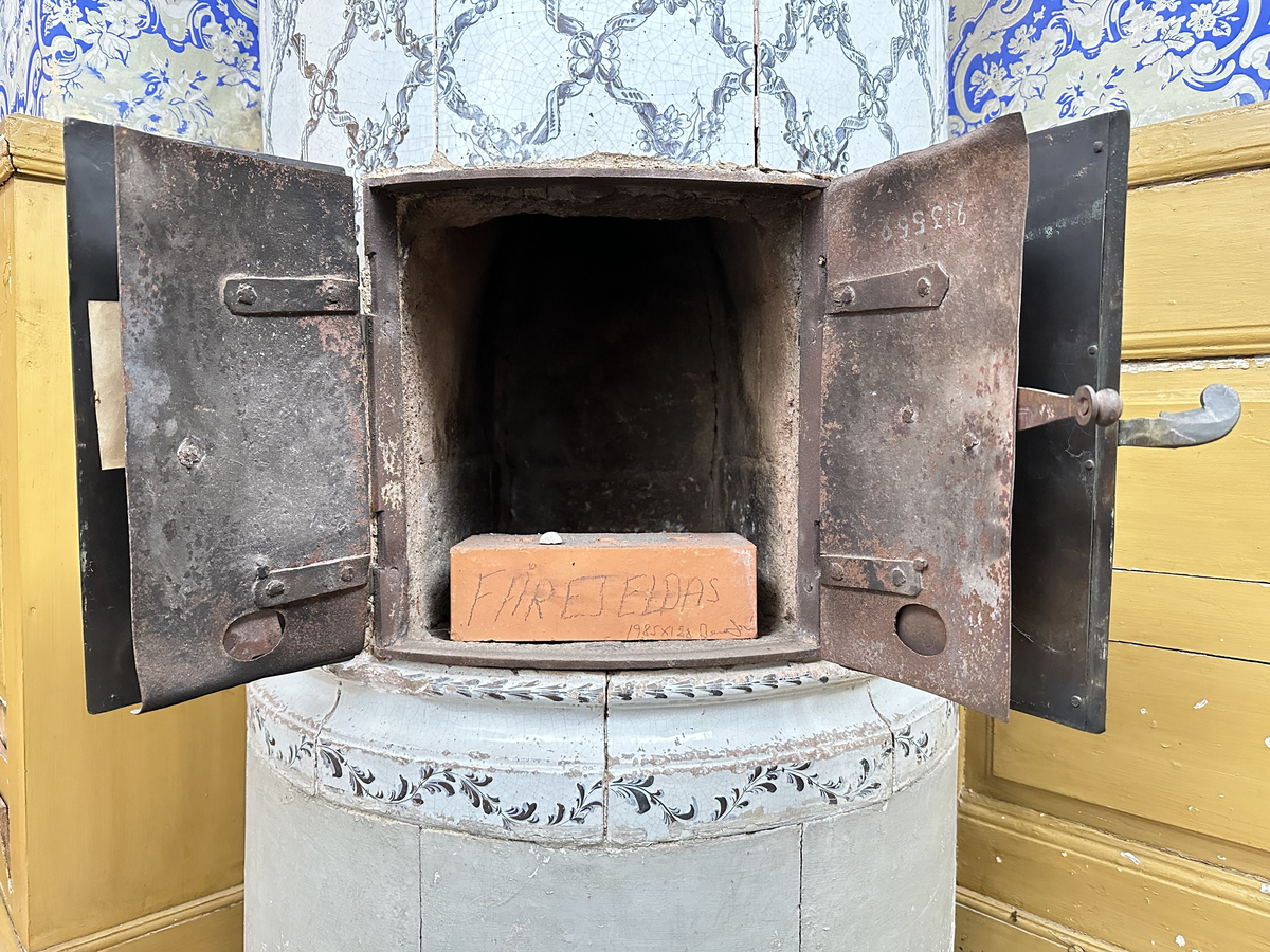 Kakelugn, rund. Klädd i kvadratiskt vitglaserat kakel, handmålat med korsande blomstergirlander i duvblått och mörkblåa detaljer. Slät sockel, gråkalkad. Fot- och översims med växlande konvex och konkav profilering samt liggande blomstergirlander. Ytterluckor i mässing med dekorativt utformade gångjärn samt klinka, i form av en sädeskärve med blommor. Innerluckor av järn, rostiga, med klinka samt två päronformade ventilluckor. Två runda sotluckor i mässing på ugnens sidor ovanför sockeln, samt en på ugnens topp. Spjällarm med ringformat handtag av järn.