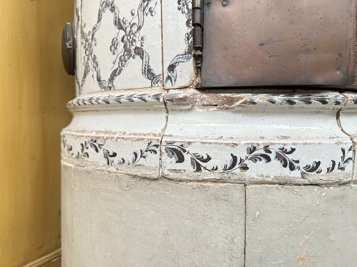 Kakelugn, rund. Klädd i kvadratiskt vitglaserat kakel, handmålat med korsande blomstergirlander i duvblått och mörkblåa detaljer. Slät sockel, gråkalkad. Fot- och översims med växlande konvex och konkav profilering samt liggande blomstergirlander. Ytterluckor i mässing med dekorativt utformade gångjärn samt klinka, i form av en sädeskärve med blommor. Innerluckor av järn, rostiga, med klinka samt två päronformade ventilluckor. Två runda sotluckor i mässing på ugnens sidor ovanför sockeln, samt en på ugnens topp. Spjällarm med ringformat handtag av järn.