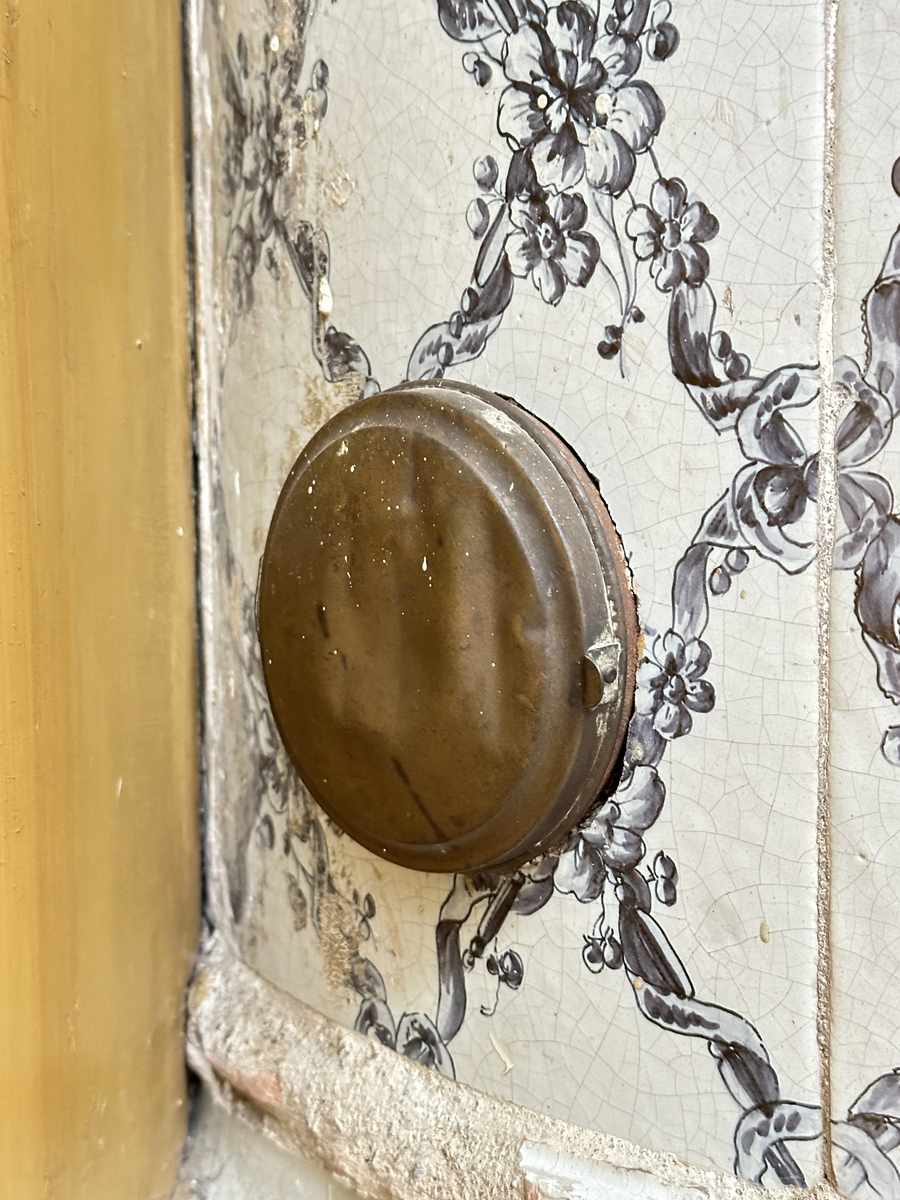 Kakelugn, rund. Klädd i kvadratiskt vitglaserat kakel, handmålat med korsande blomstergirlander i duvblått och mörkblåa detaljer. Slät sockel, gråkalkad. Fot- och översims med växlande konvex och konkav profilering samt liggande blomstergirlander. Ytterluckor i mässing med dekorativt utformade gångjärn samt klinka, i form av en sädeskärve med blommor. Innerluckor av järn, rostiga, med klinka samt två päronformade ventilluckor. Två runda sotluckor i mässing på ugnens sidor ovanför sockeln, samt en på ugnens topp. Spjällarm med ringformat handtag av järn.