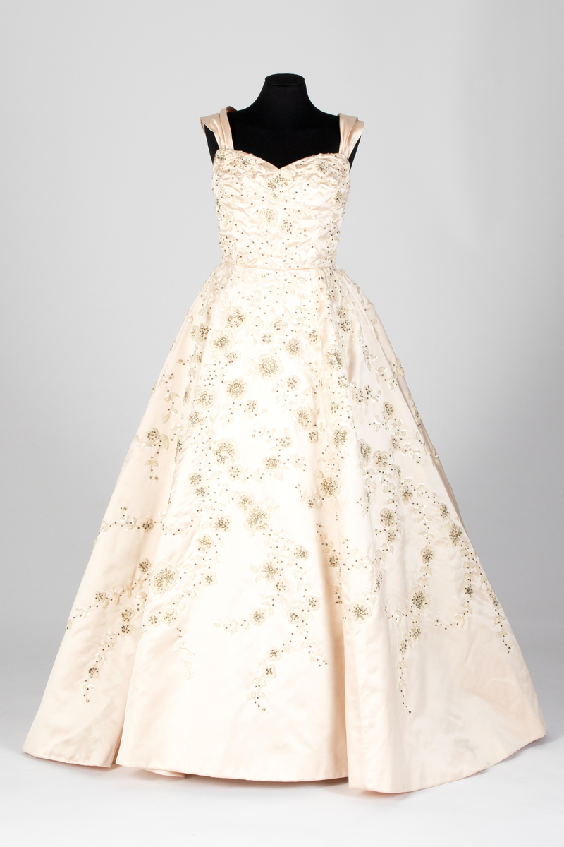 Fotsid ballkjole av lys rosa, satengvevd duchesse silke. Kjolen er delt i midten, har figursydd liv med spiler, hjerteformet avstivet utringning og glidelås i ryggen. Brede skulderbånd som kan legges ned fra skuldrene. Liver er fôret med hvitt bomullsstoff. Støttebånd under bysten. Skjørtet er sterkt utskrådd, av sju bredder og er fôret med tynt, stivt syntetisk stoff. Kjolen er brodert med store blomsterranker i benhvit chenille, hvite paljetter, perler og mørke stener. Med kjolen følger et voluminøst underskjørt av flere lag stivt nettstoff, underst et taftskjørt, fôret med nettstoff.