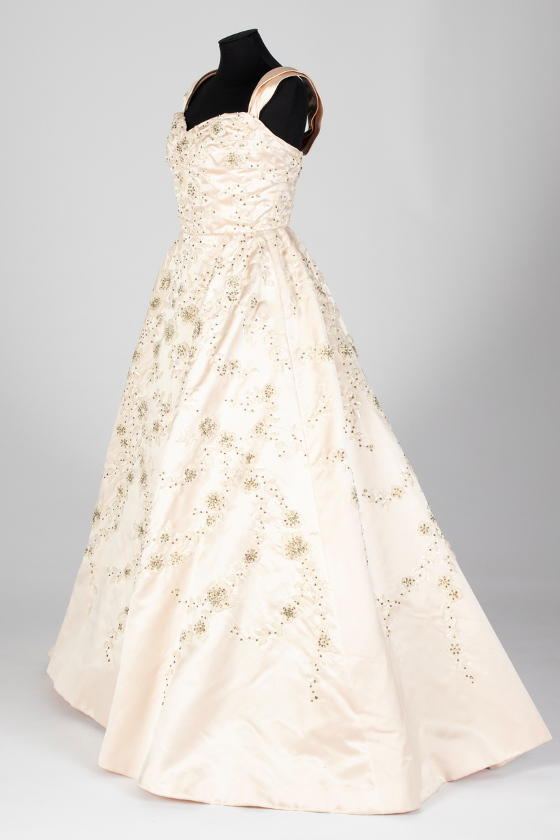 Fotsid ballkjole av lys rosa, satengvevd duchesse silke. Kjolen er delt i midten, har figursydd liv med spiler, hjerteformet avstivet utringning og glidelås i ryggen. Brede skulderbånd som kan legges ned fra skuldrene. Liver er fôret med hvitt bomullsstoff. Støttebånd under bysten. Skjørtet er sterkt utskrådd, av sju bredder og er fôret med tynt, stivt syntetisk stoff. Kjolen er brodert med store blomsterranker i benhvit chenille, hvite paljetter, perler og mørke stener. Med kjolen følger et voluminøst underskjørt av flere lag stivt nettstoff, underst et taftskjørt, fôret med nettstoff.