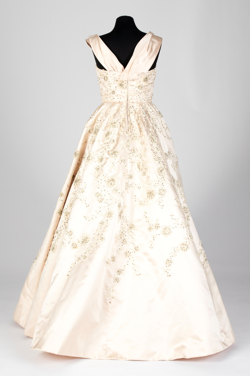 Fotsid ballkjole av lys rosa, satengvevd duchesse silke. Kjolen er delt i midten, har figursydd liv med spiler, hjerteformet avstivet utringning og glidelås i ryggen. Brede skulderbånd som kan legges ned fra skuldrene. Liver er fôret med hvitt bomullsstoff. Støttebånd under bysten. Skjørtet er sterkt utskrådd, av sju bredder og er fôret med tynt, stivt syntetisk stoff. Kjolen er brodert med store blomsterranker i benhvit chenille, hvite paljetter, perler og mørke stener. Med kjolen følger et voluminøst underskjørt av flere lag stivt nettstoff, underst et taftskjørt, fôret med nettstoff.