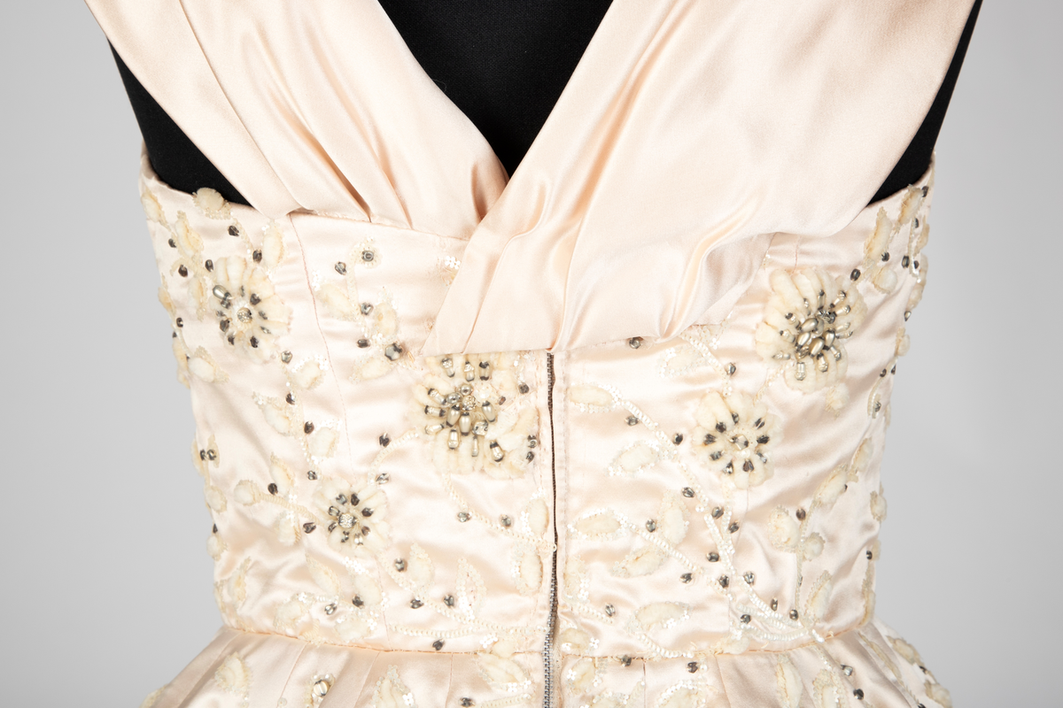 Fotsid ballkjole av lys rosa, satengvevd duchesse silke. Kjolen er delt i midten, har figursydd liv med spiler, hjerteformet avstivet utringning og glidelås i ryggen. Brede skulderbånd som kan legges ned fra skuldrene. Liver er fôret med hvitt bomullsstoff. Støttebånd under bysten. Skjørtet er sterkt utskrådd, av sju bredder og er fôret med tynt, stivt syntetisk stoff. Kjolen er brodert med store blomsterranker i benhvit chenille, hvite paljetter, perler og mørke stener. Med kjolen følger et voluminøst underskjørt av flere lag stivt nettstoff, underst et taftskjørt, fôret med nettstoff.