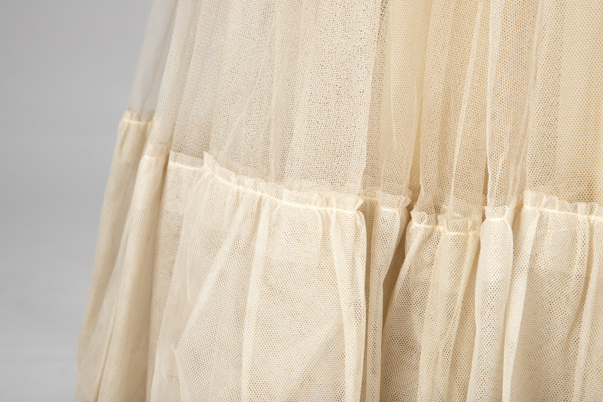 Fotsid ballkjole av lys rosa, satengvevd duchesse silke. Kjolen er delt i midten, har figursydd liv med spiler, hjerteformet avstivet utringning og glidelås i ryggen. Brede skulderbånd som kan legges ned fra skuldrene. Liver er fôret med hvitt bomullsstoff. Støttebånd under bysten. Skjørtet er sterkt utskrådd, av sju bredder og er fôret med tynt, stivt syntetisk stoff. Kjolen er brodert med store blomsterranker i benhvit chenille, hvite paljetter, perler og mørke stener. Med kjolen følger et voluminøst underskjørt av flere lag stivt nettstoff, underst et taftskjørt, fôret med nettstoff.