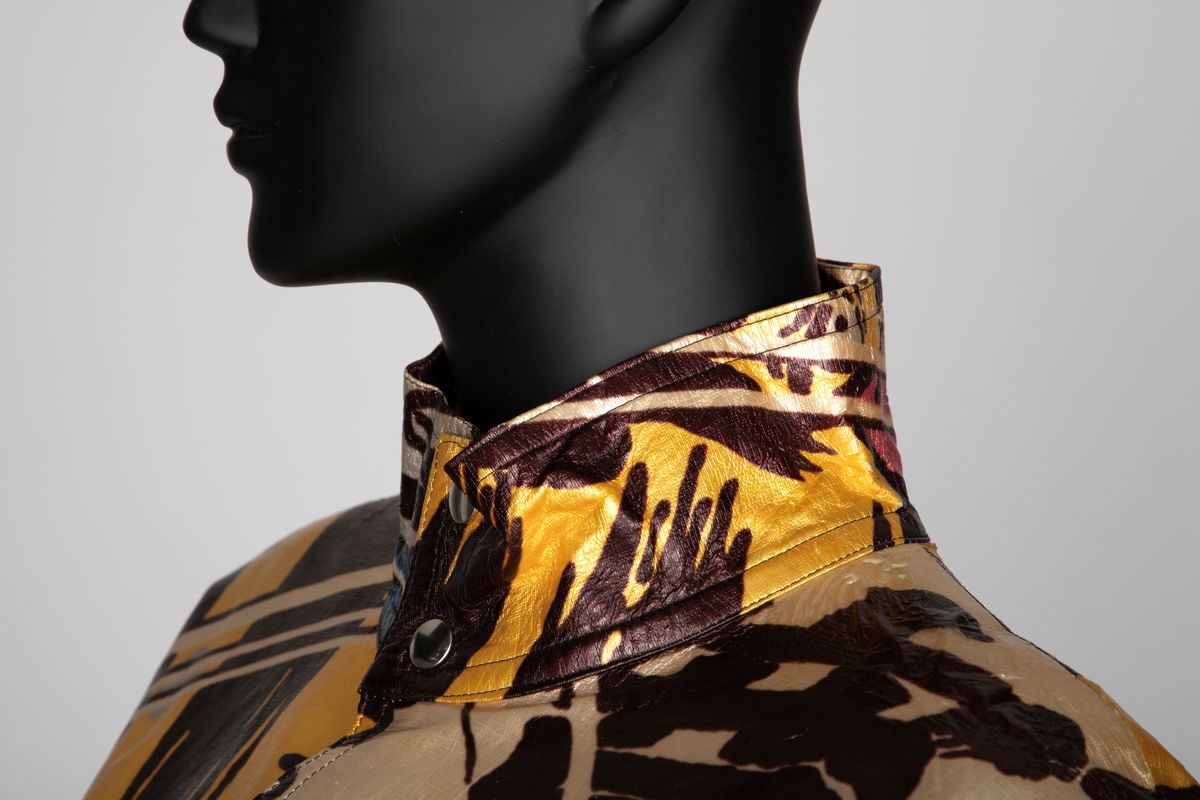 Jakke av vinyl i fargene rosa, blå, brun, rød og gul med motiv av samurai i tegneseriestil. Jakken er romslig, har rette linjer, nedsenket skulder, dypt ermhull og høy halslinning. Lukkes med trykknapper. Fôret med svart stoff.