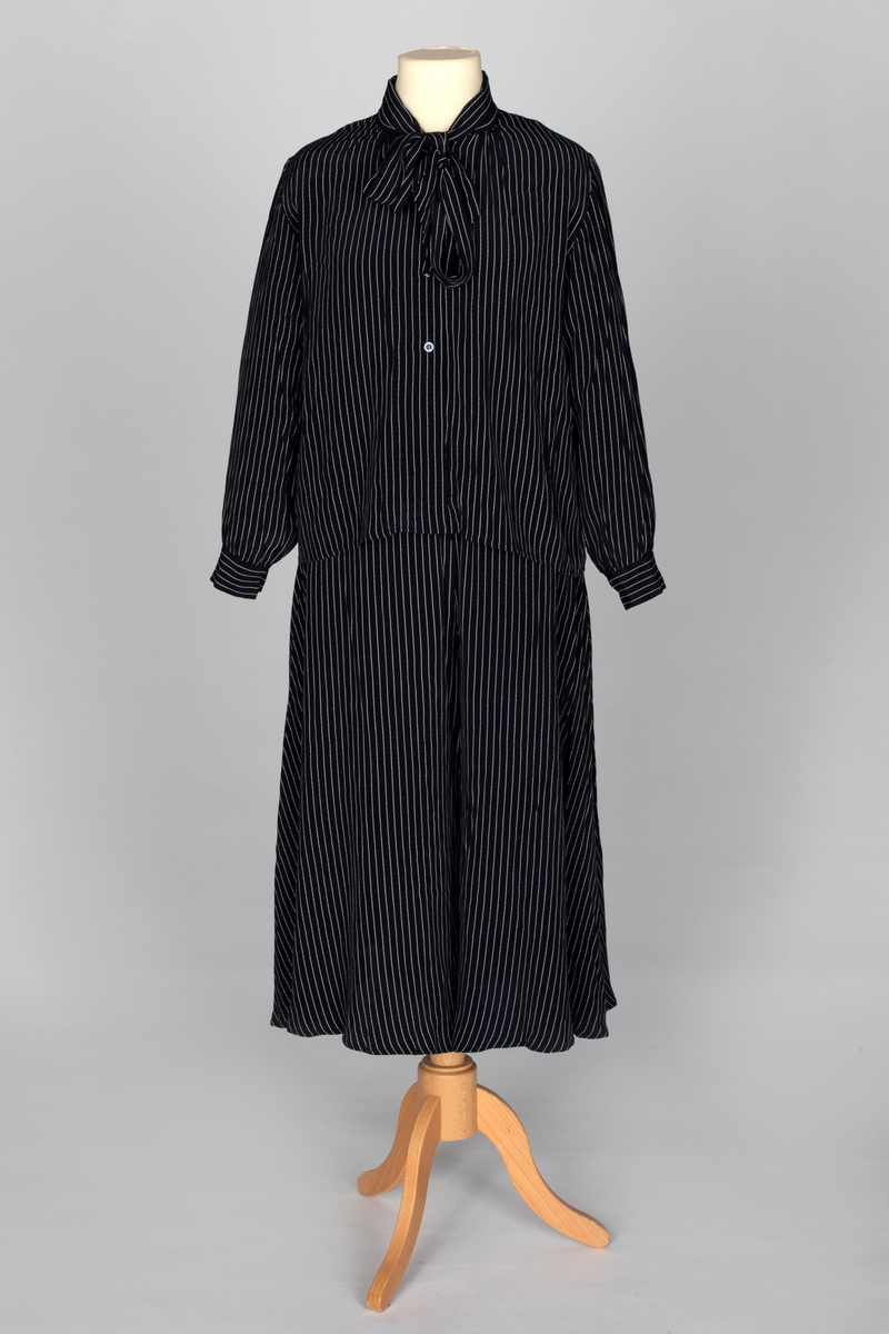 Todelt drakt i mørkeblå viskose med lyse striper, bestående av et bukseskjørt og en bluse, begge med god vidde. Blusen har lange ermer med mansjetter, knepping midt foran og et fastsydd skjerf som kan knyttes til en sløyfe. Bukseskjørtet har smal linning og glidelås på venstre side.