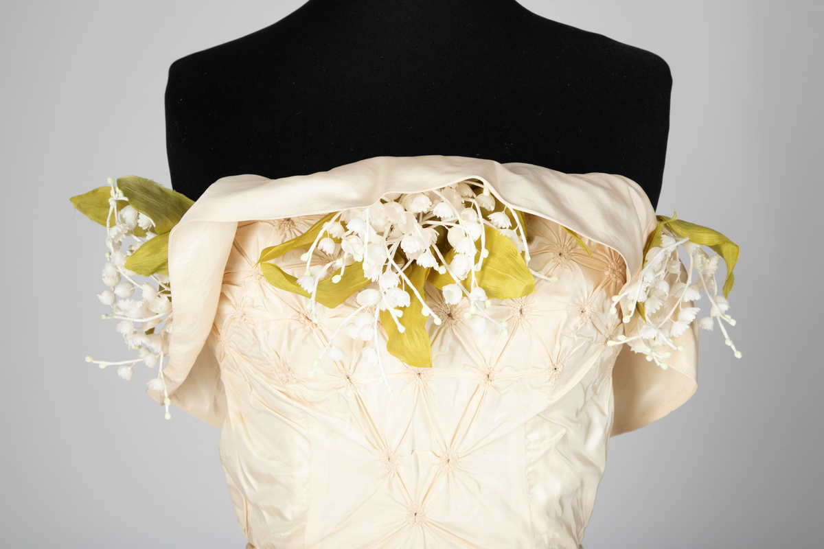 Fotsid ball-/ brudekjole i kremhvit silke med lite slep. Tettsittende, stroppeløs overdel med volangkappe øverst. Overdelen og øvre del av skjørtet består av håndsydde, "snurpete" rosetter. Skjørtet går ned i spiss foran, på sidene og bak. Buketter av liljekonvaller i plastmateriale er festet langs overdelens volangkappe.