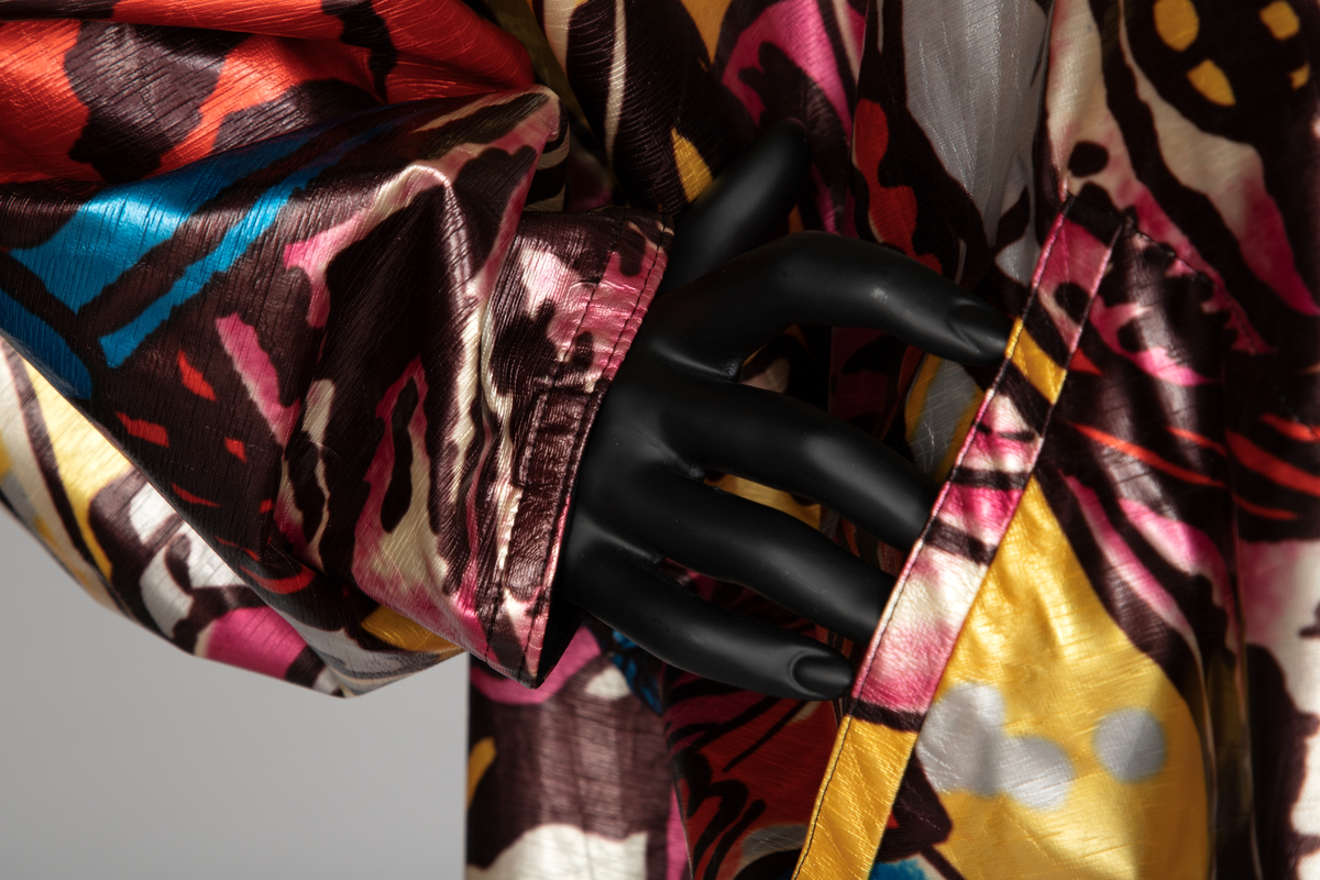 Jakke av vinyl i fargene rosa, blå, brun, rød og gul med motiv av samurai i tegneseriestil. Jakken er romslig, har rette linjer, nedsenket skulder, dypt ermhull og høy halslinning. Lukkes med trykknapper. Fôret med svart stoff.