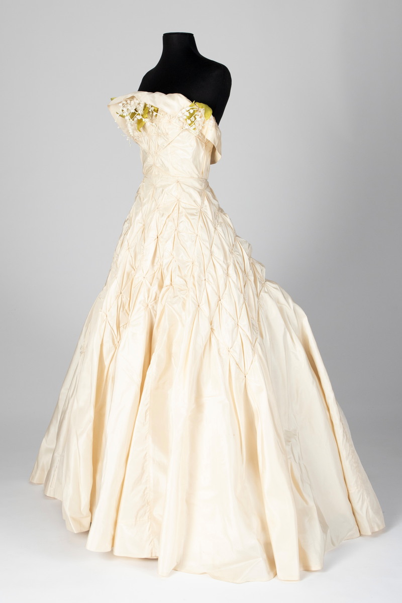 Fotsid ball-/ brudekjole i kremhvit silke med lite slep. Tettsittende, stroppeløs overdel med volangkappe øverst. Overdelen og øvre del av skjørtet består av håndsydde, "snurpete" rosetter. Skjørtet går ned i spiss foran, på sidene og bak. Buketter av liljekonvaller i plastmateriale er festet langs overdelens volangkappe.