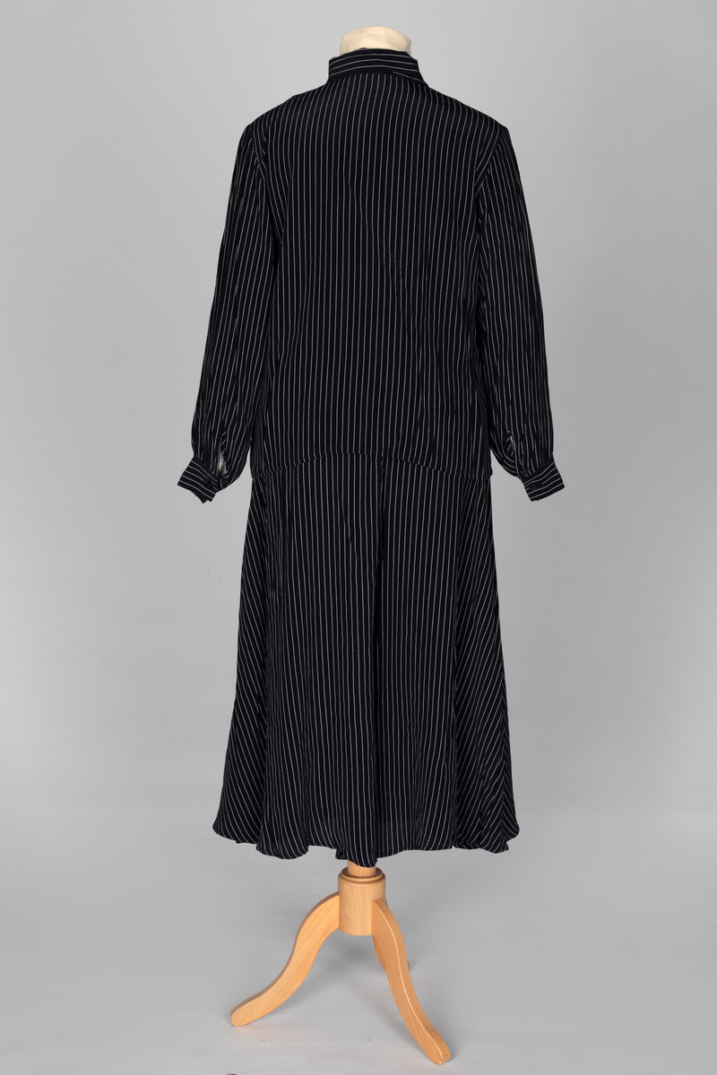 Todelt drakt i mørkeblå viskose med lyse striper, bestående av et bukseskjørt og en bluse, begge med god vidde. Blusen har lange ermer med mansjetter, knepping midt foran og et fastsydd skjerf som kan knyttes til en sløyfe. Bukseskjørtet har smal linning og glidelås på venstre side.