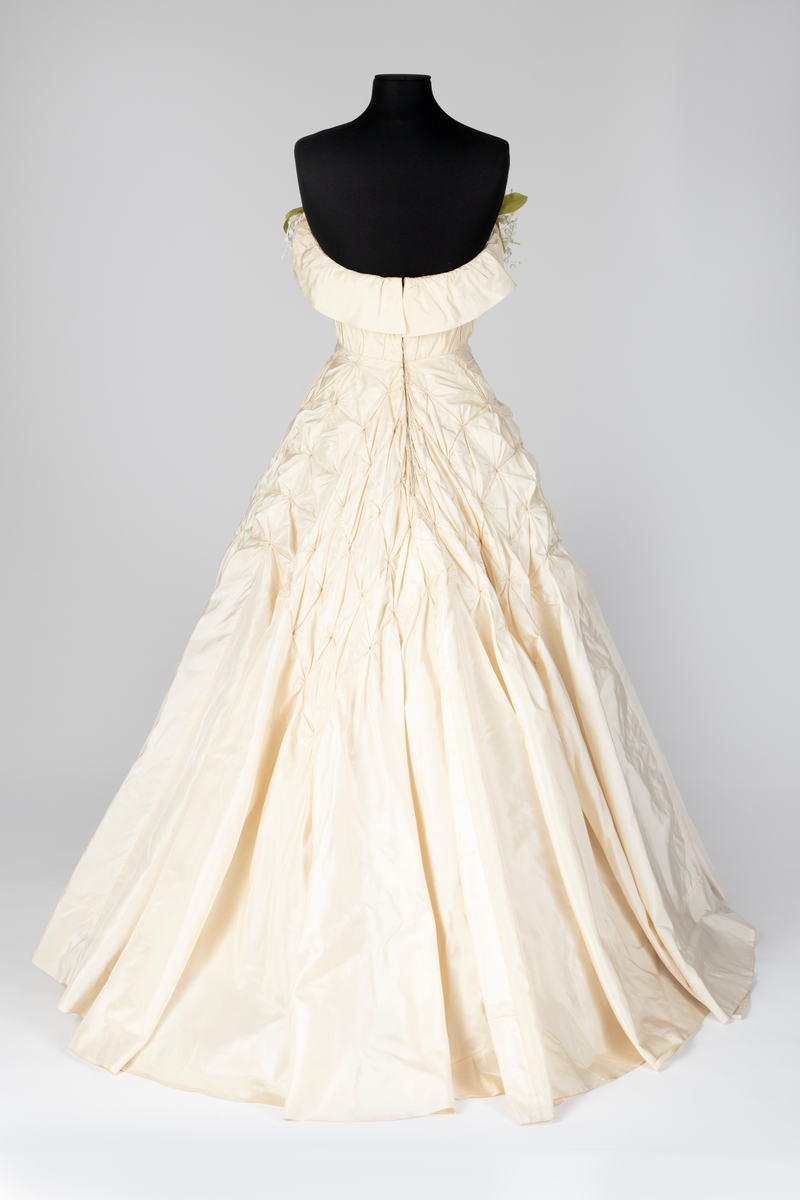 Fotsid ball-/ brudekjole i kremhvit silke med lite slep. Tettsittende, stroppeløs overdel med volangkappe øverst. Overdelen og øvre del av skjørtet består av håndsydde, "snurpete" rosetter. Skjørtet går ned i spiss foran, på sidene og bak. Buketter av liljekonvaller i plastmateriale er festet langs overdelens volangkappe.