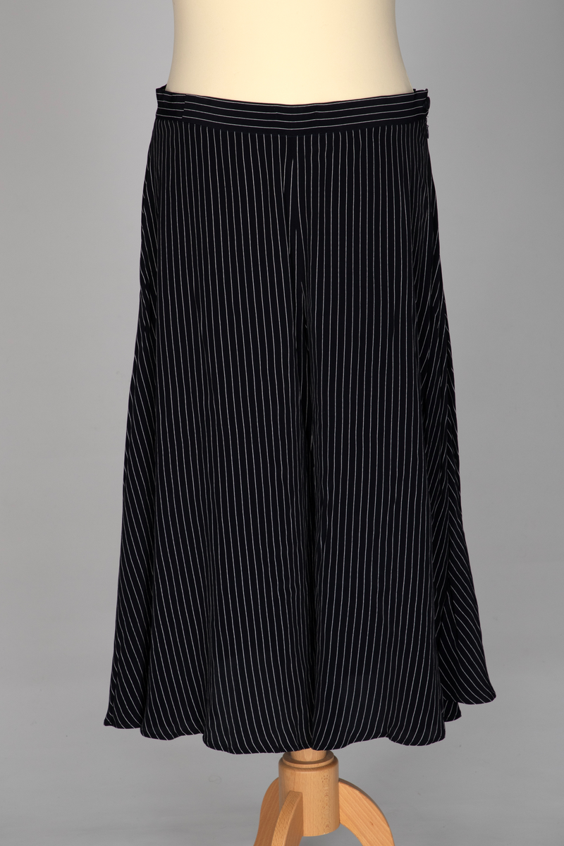 Todelt drakt i mørkeblå viskose med lyse striper, bestående av et bukseskjørt og en bluse, begge med god vidde. Blusen har lange ermer med mansjetter, knepping midt foran og et fastsydd skjerf som kan knyttes til en sløyfe. Bukseskjørtet har smal linning og glidelås på venstre side.