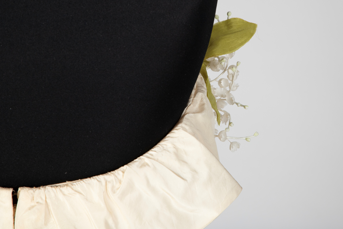 Fotsid ball-/ brudekjole i kremhvit silke med lite slep. Tettsittende, stroppeløs overdel med volangkappe øverst. Overdelen og øvre del av skjørtet består av håndsydde, "snurpete" rosetter. Skjørtet går ned i spiss foran, på sidene og bak. Buketter av liljekonvaller i plastmateriale er festet langs overdelens volangkappe.