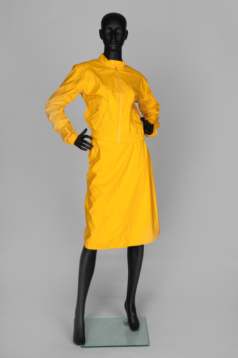 Drakt som består av battlejakke og omslagsskjørt i gult regntøy. Jakken har frontlukning med enveisglidelås og linning i halsen lukket med trykknapp. Skråstilt lomme, lukket med glidelås nederst på hvert forstykke. Jakken har innsydd strikk i nedre kant og isydde, tosøms-ermer og bærestykke. Ermer og bole er avsluttet med isydd elastikk i nederkant. Dette gir en romslig blusong-effekt. Alle viktige sømmer er både sydd og sveiset. Knehøyt omslagsskjørt med åpning midt bak, isydd strikk i livet og knepping med to trykknapper der skjørtet har sin circa 10cm's overlapping bak. Skjørtet er klippet i ett stykke og kantet i nederkant med påsydd skråbånd.