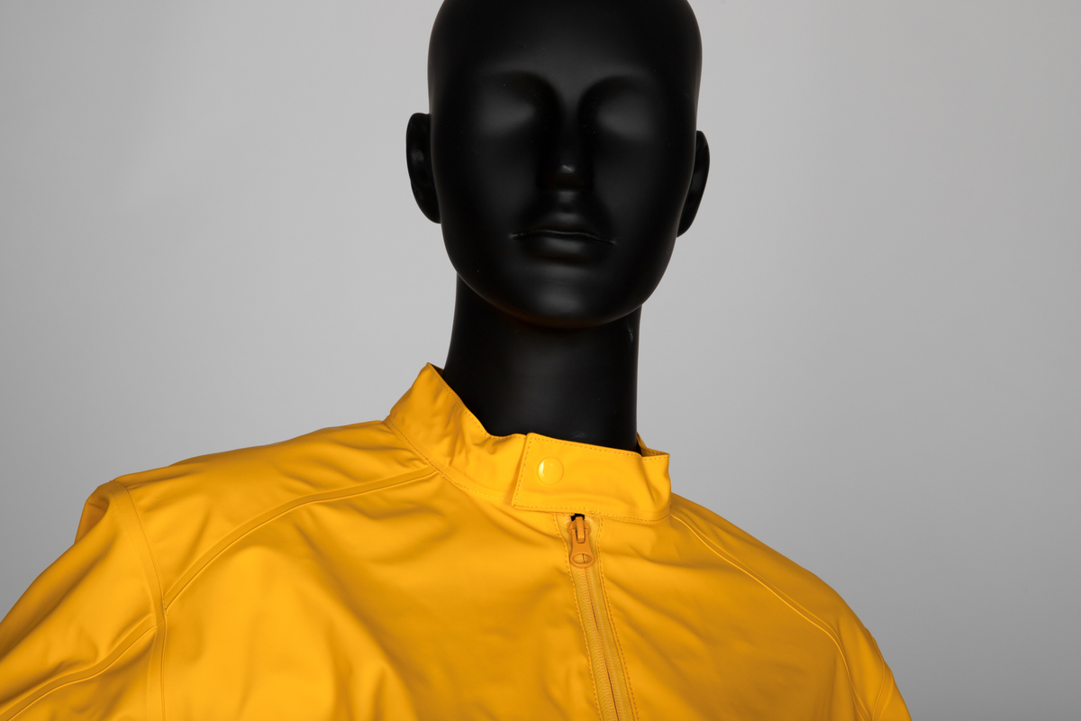 Drakt som består av battlejakke og omslagsskjørt i gult regntøy. Jakken har frontlukning med enveisglidelås og linning i halsen lukket med trykknapp. Skråstilt lomme, lukket med glidelås nederst på hvert forstykke. Jakken har innsydd strikk i nedre kant og isydde, tosøms-ermer og bærestykke. Ermer og bole er avsluttet med isydd elastikk i nederkant. Dette gir en romslig blusong-effekt. Alle viktige sømmer er både sydd og sveiset. Knehøyt omslagsskjørt med åpning midt bak, isydd strikk i livet og knepping med to trykknapper der skjørtet har sin circa 10cm's overlapping bak. Skjørtet er klippet i ett stykke og kantet i nederkant med påsydd skråbånd.